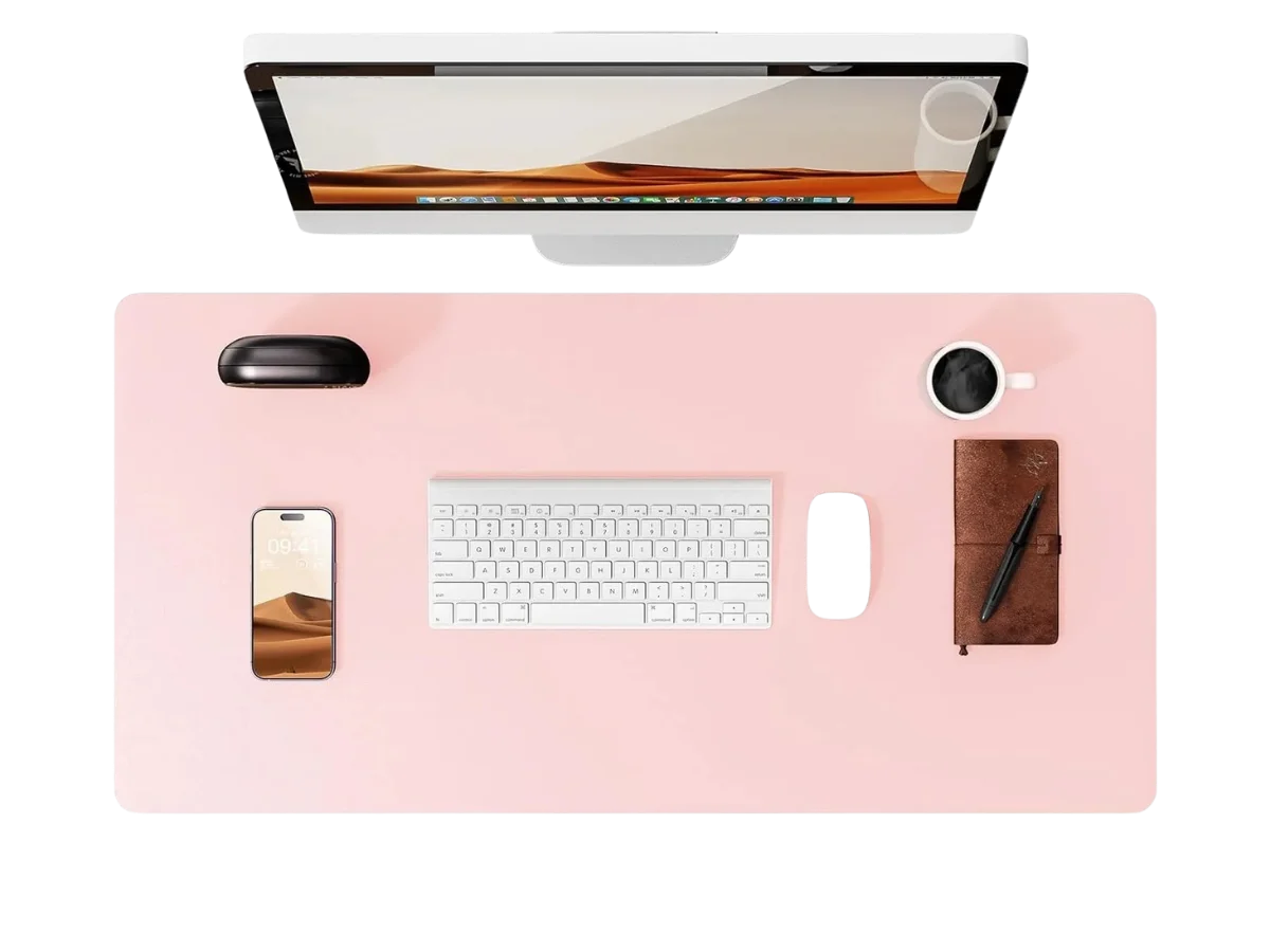 Vue de dessus d'un bureau organisé avec un grand tapis de souris cuir rose pastel, un écran d'ordinateur, un clavier, une souris noire, un smartphone, une tasse de café et un carnet en cuir.