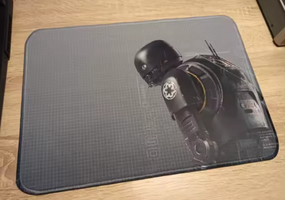 Un tapis de souris gris foncé avec le droïde Star Wars K-2SO de Rogue One imprimé dessus, posé sur une surface en bois.