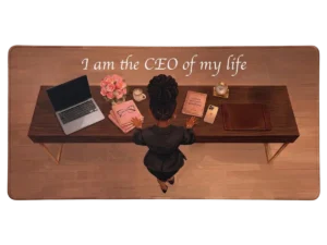 Vue de dessus d'une femme noire assise à un bureau, ce tapis de souris rose contient l'inscription 'I am the CEO of my life'.